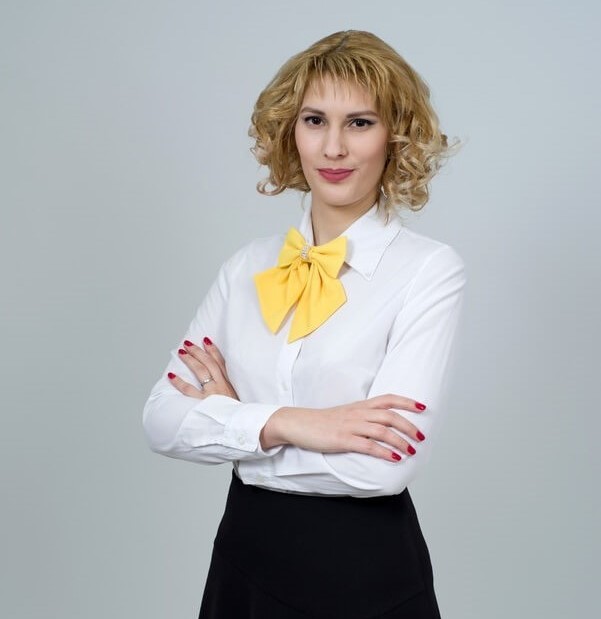 Professional woman with curly blonde hair, wearing a white blouse and a yellow bow tie, arms crossed, against a neutral gray background, representing transparency in crypto gambling at InstantCryptoWins.com.