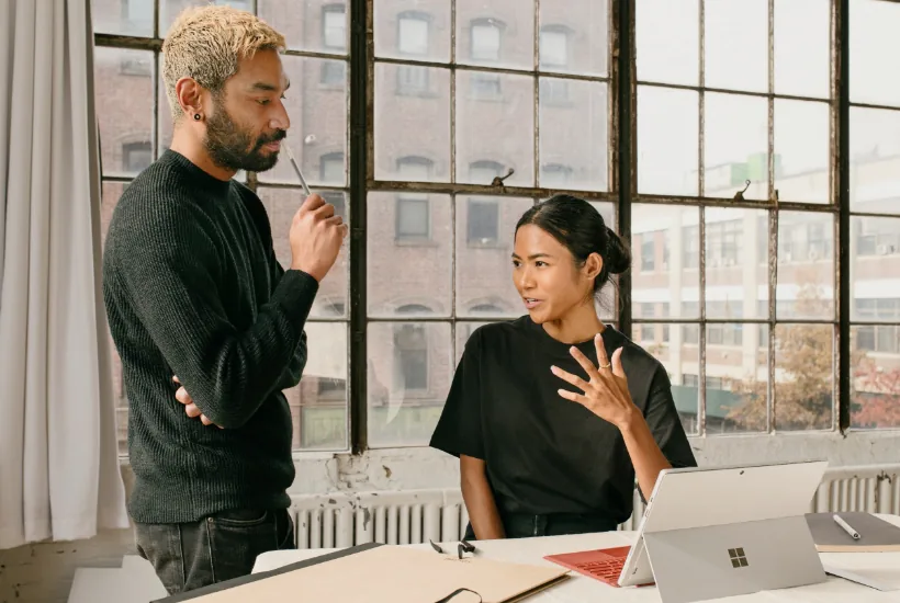 Two individuals engaged in a discussion in a modern office setting, with one person holding a pen and the other gesturing, while a laptop sits on the table, reflecting a collaborative atmosphere related to cryptocurrency strategies.