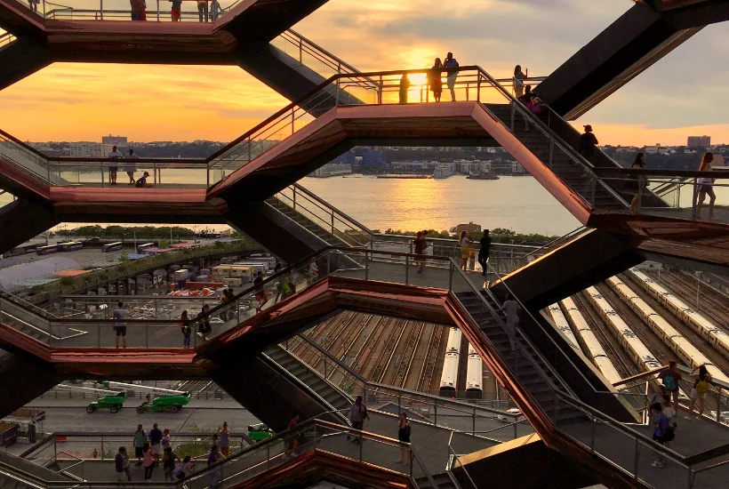 Sunset view of a modern architectural structure with multiple levels, people walking and gathering on staircases, overlooking a river and city skyline, reflecting themes of urban exploration and engagement.